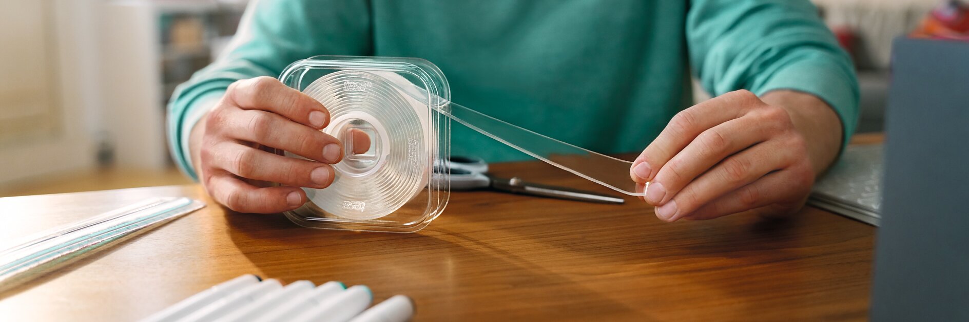 A person wearing a green shirt is seated at a wooden table, handling a clear tesa tape dispenser. Several markers are scattered on the table, and a notebook is partially visible. Scissors are also present near the persons hands. (This text has been generated by AI)