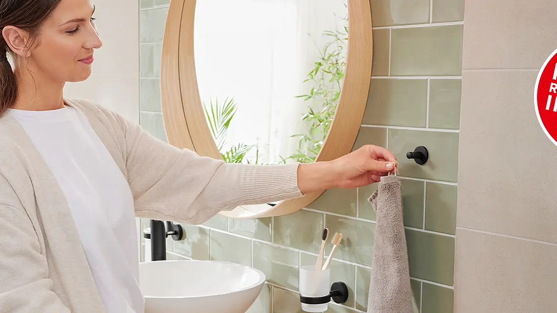 A woman stands in a bathroom setting, smiling and hanging a hand towel on a black hook with tesa tape. In the background, there is a circular mirror, a round sink, and some toiletries. A red circular sign reads, Find the right product in 2 min. (This text has been generated by AI)