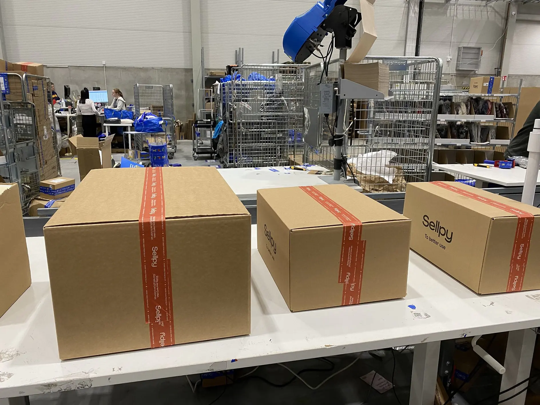 Kolm pappkarpi, millel on punane *tesa teip* ja silt "Sellpy", on asetatud valgele lauale laos. Taustal on näha riiuleid, pakke ja töötajaid, kes tegelevad sorteerimise ja pakkimisega.