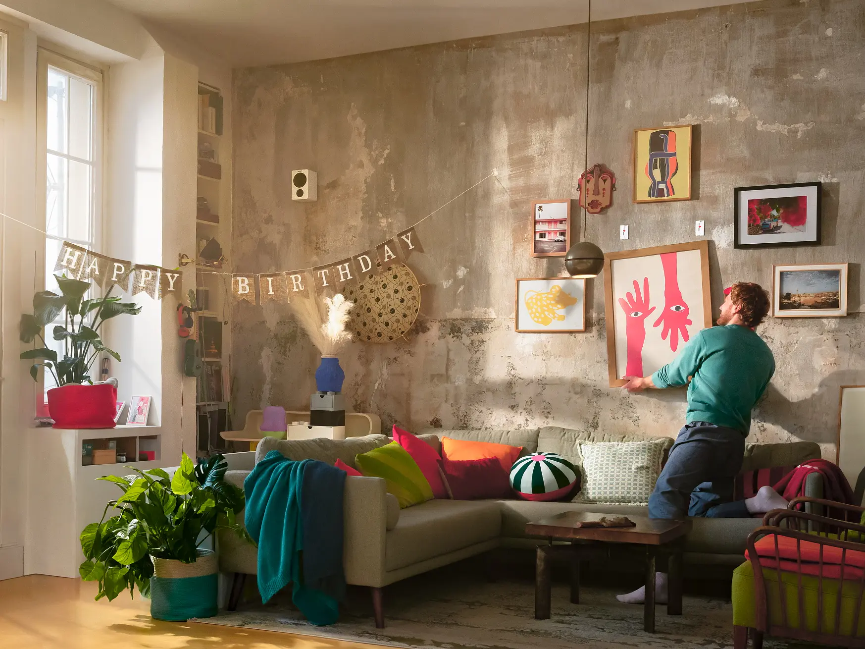 Inimene riputab raamitud kunstiteose tekstuurse seinaga elutoas, mis on sisustatud taimede, raamaturiiulite ja värviliste patjadega. (This text has been generated by AI)