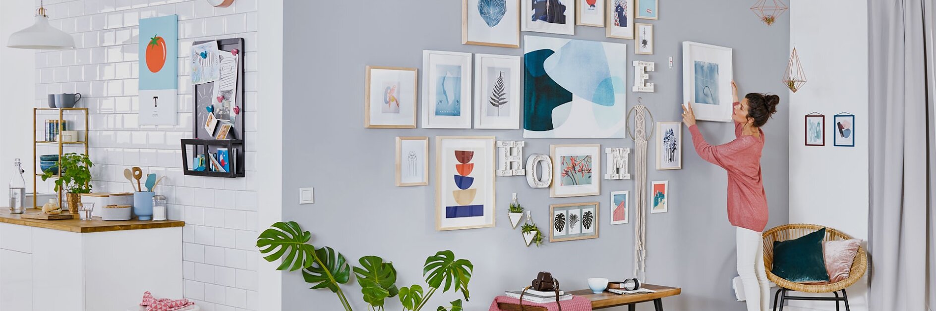 A woman in a pink shirt is arranging frames on a grey and white wall, filled with various artwork and photos, using tesa tape. The room has a modern design with a small kitchen on the left, light wood flooring, and green plants for decoration. (This text has been generated by AI)