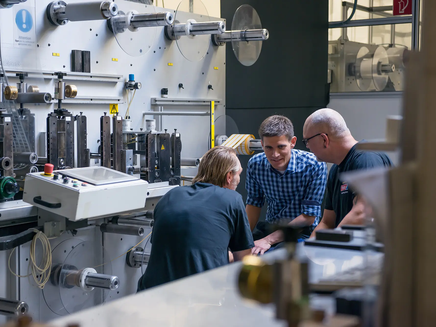 Three men are gathered around a large industrial machine in a factory setting. They appear to be engaged in discussion. The machine has various levers and components, and there are rolls of tesa tape in the background. (This text has been generated by AI)