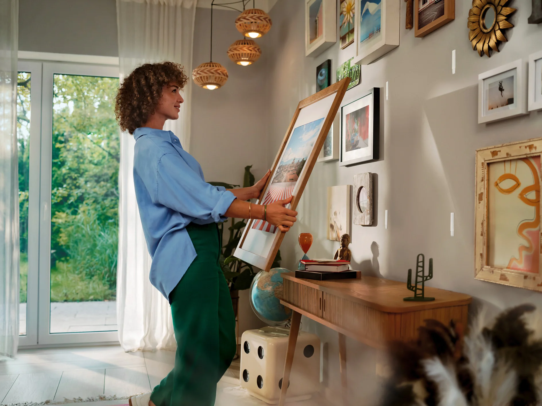 A person in a blue shirt and green pants is hanging a framed picture on a wall using tesa tape. The room is decorated with multiple artworks and has a wooden desk with various items. A window with sheer curtains is open to a view of greenery outside. (This text has been generated by AI)