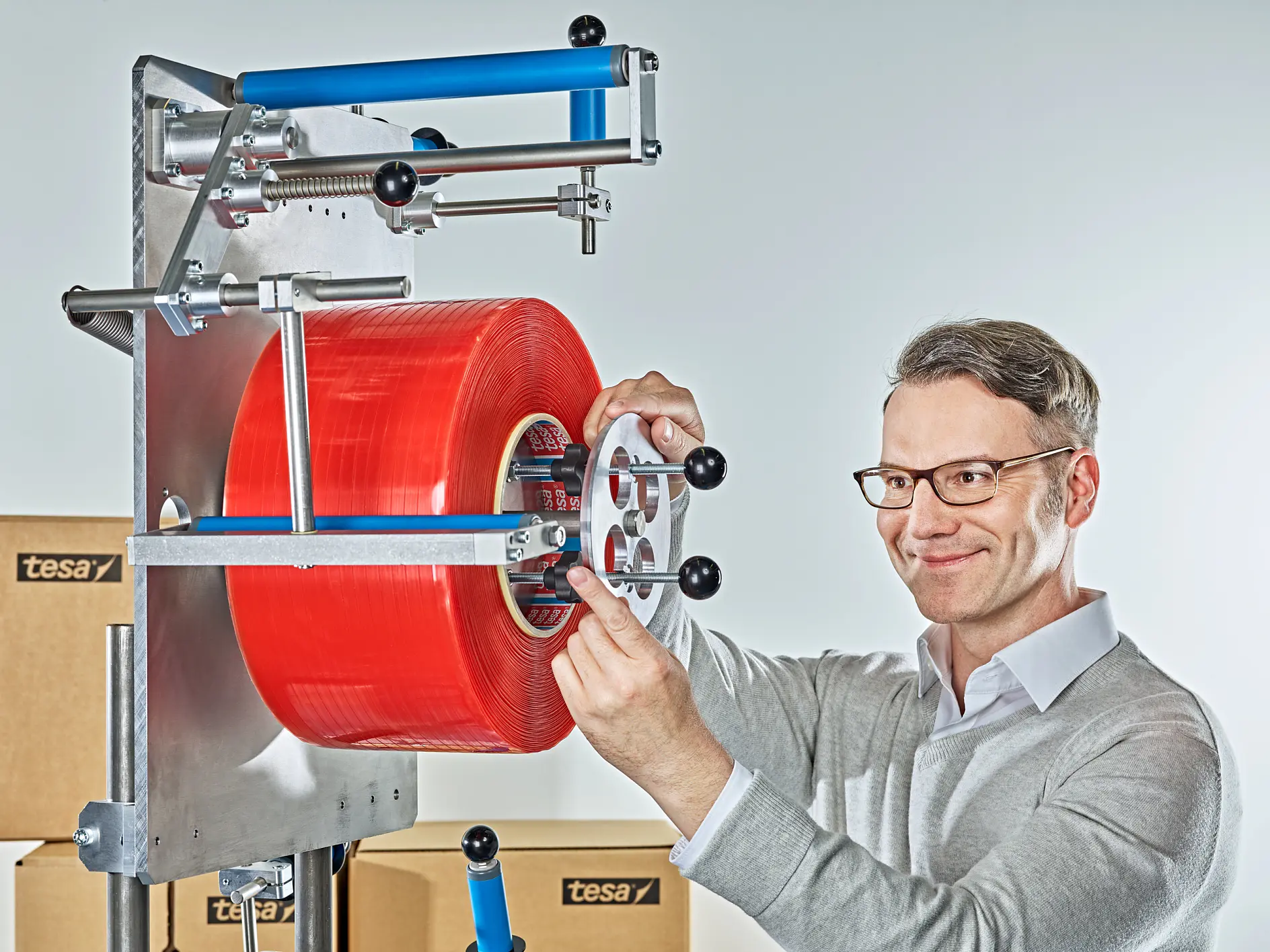 man adjusting red packaging tape on industrial taping machine in warehouse with stacked boxes labeled tesa (This text has been generated by AI)