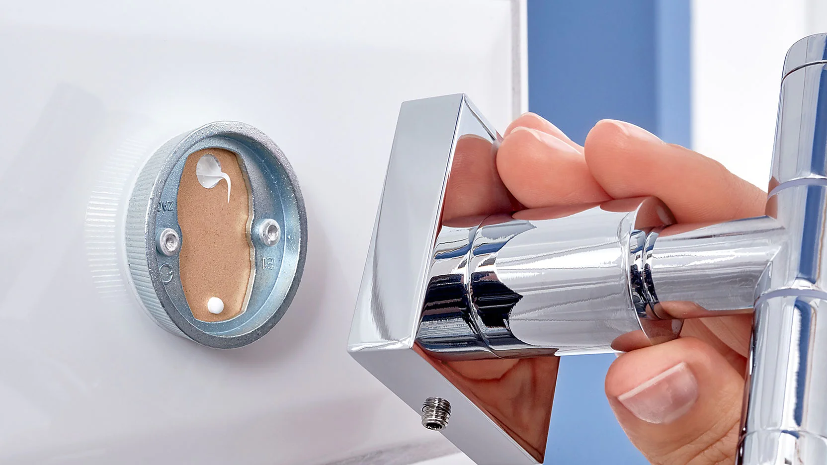 A hand is holding a shiny chrome door handle against a white door. The handle is partially detached, revealing the mounting plate and screws inside the door. The image focuses on the installation or repair process of the door handle using tesa tape. (This text has been generated by AI)