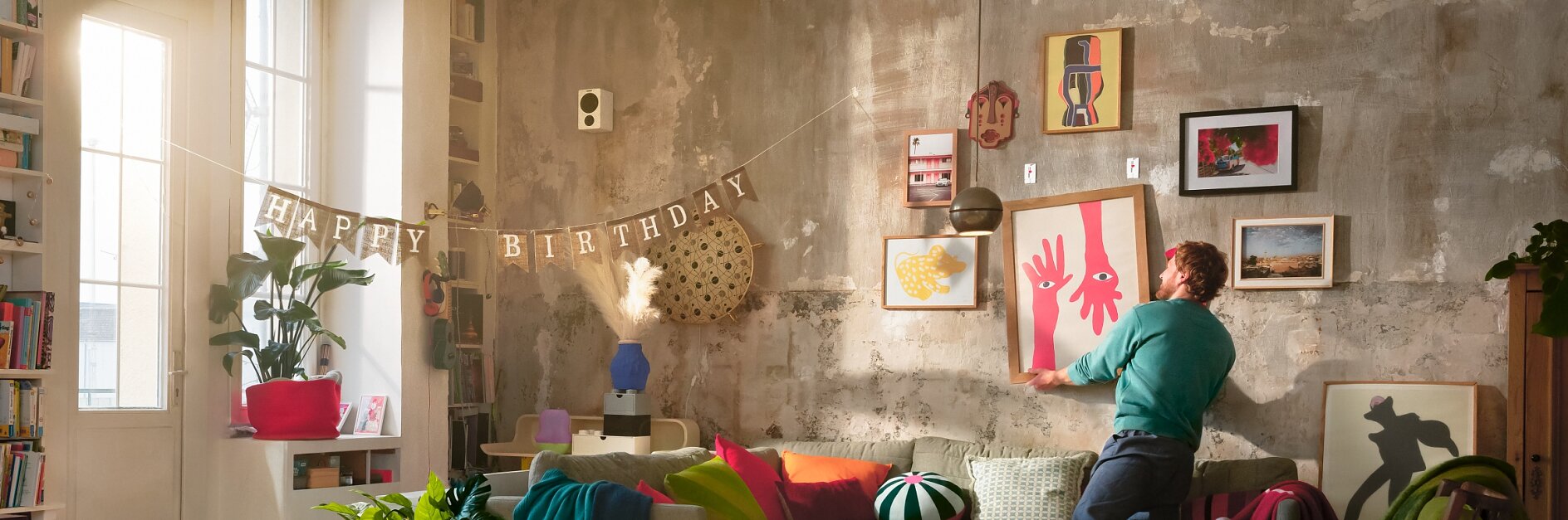 Kodikas olohuone, jossa on rouhea betoniseinä koristeltuna kehystetyillä tauluilla. Henkilö ripustaa punaista abstraktia maalausta vihreän sohvan yläpuolelle, joka on täynnä värikkäitä tyynyjä. Huoneessa on kirkasta luonnonvaloa korkeista ikkunoista, viherkasveja ja sekoitus modernia ja vintage-huonekaluja.