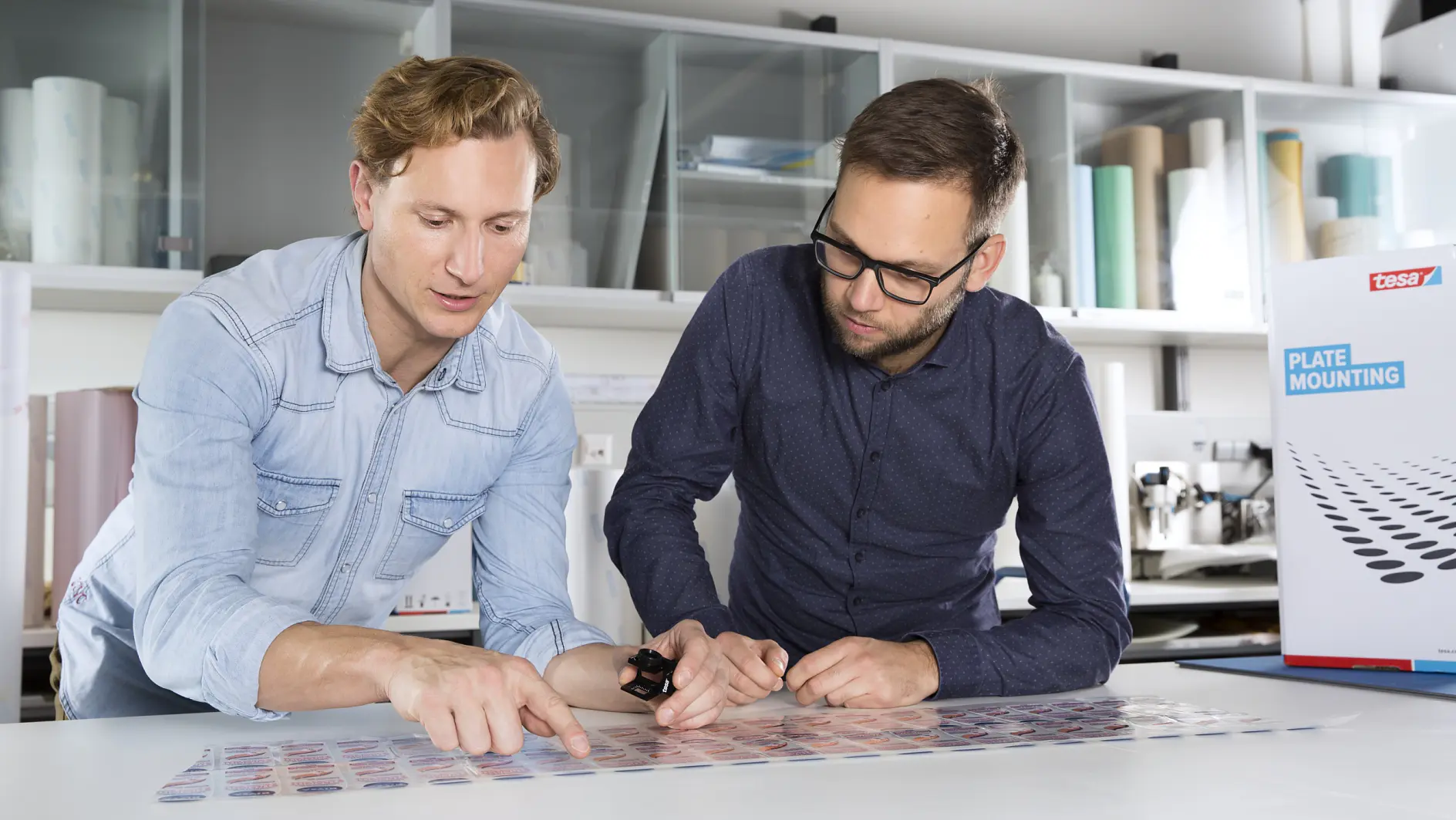 Two men are focused on examining a sheet of small printed images on a table. One is holding a magnifying tool. Behind them are shelves with rolls of tesa tape and various office supplies. A box labeled plate mounting is on the right. (This text has been generated by AI)