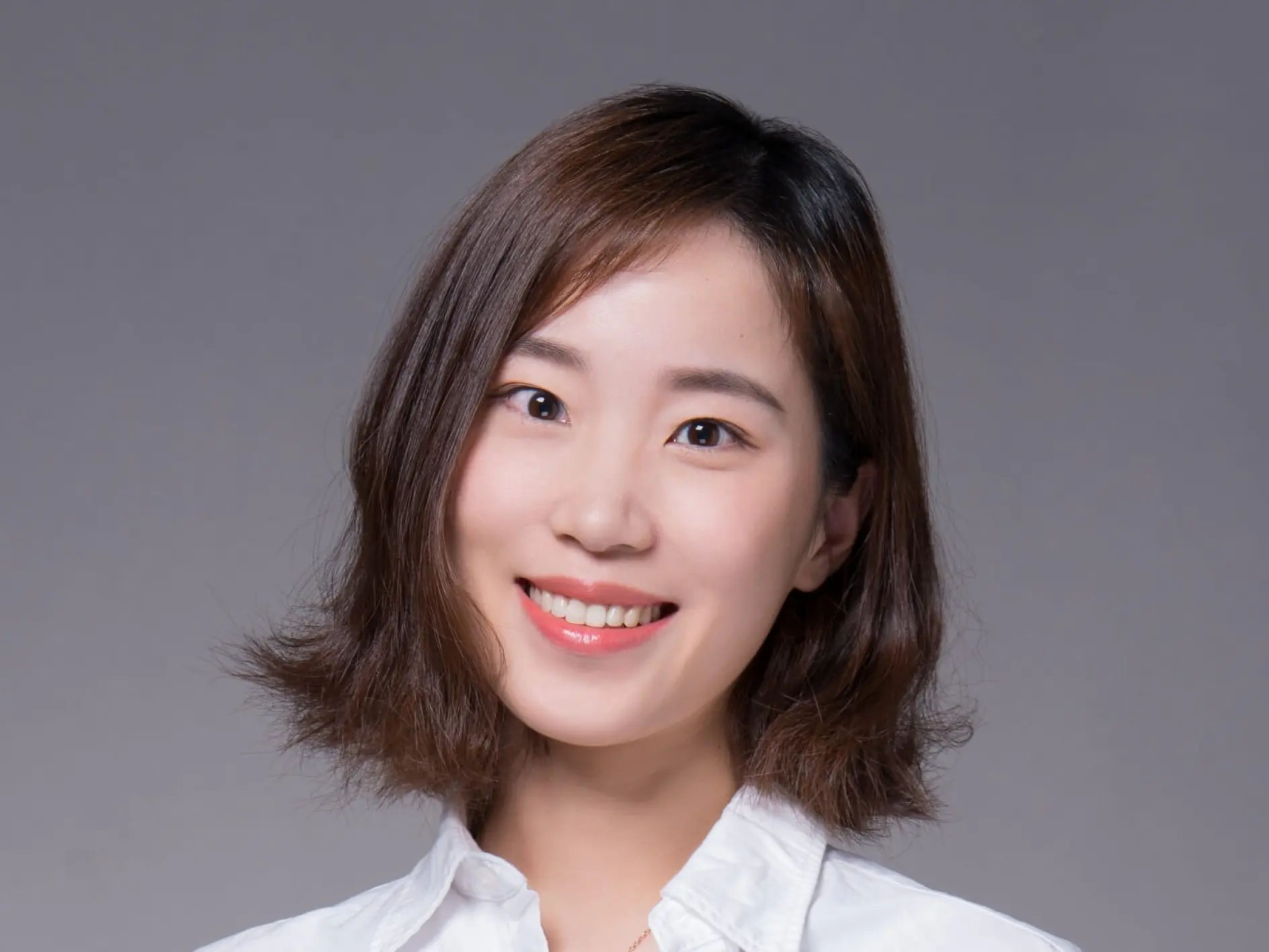 A person with short, wavy brown hair is smiling and wearing a white collared shirt. The background is gray, and the lighting is bright and even.