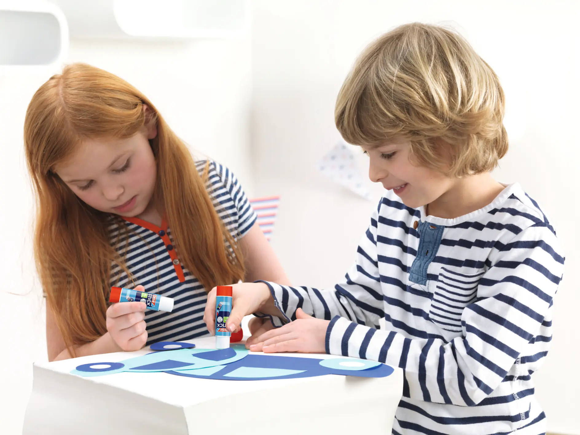 two children, a girl and a boy, sitting indoors and using glue sticks to create a craft on paper at a white table tesa (This text has been generated by AI)