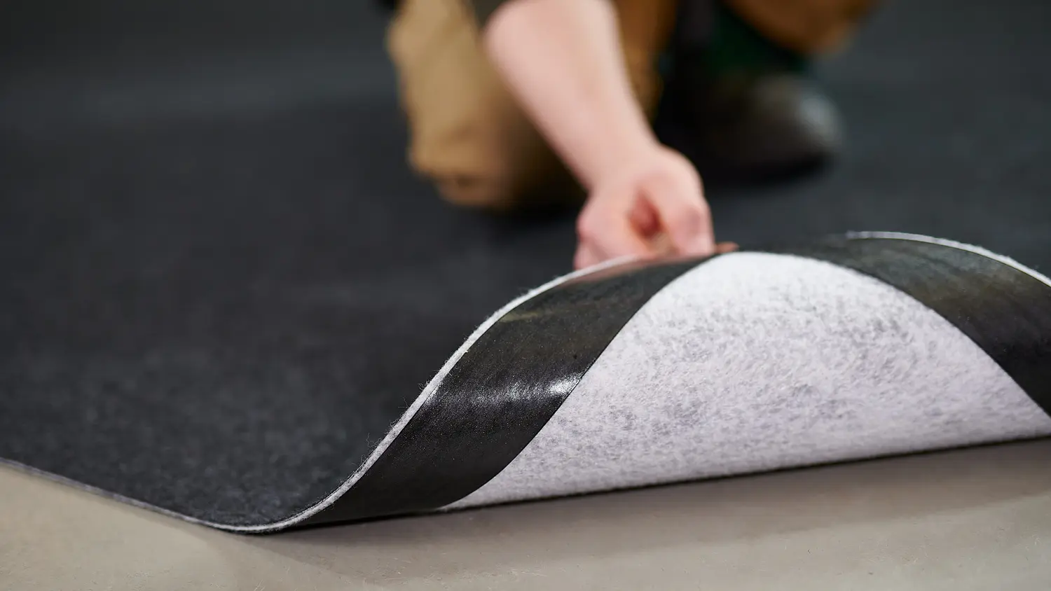 A person wearing brown pants is shown installing a black and white carpet tile over a smooth, gray floor. The focus is on the persons hand lifting the edge of the tile, revealing the underside secured with tesa tape.