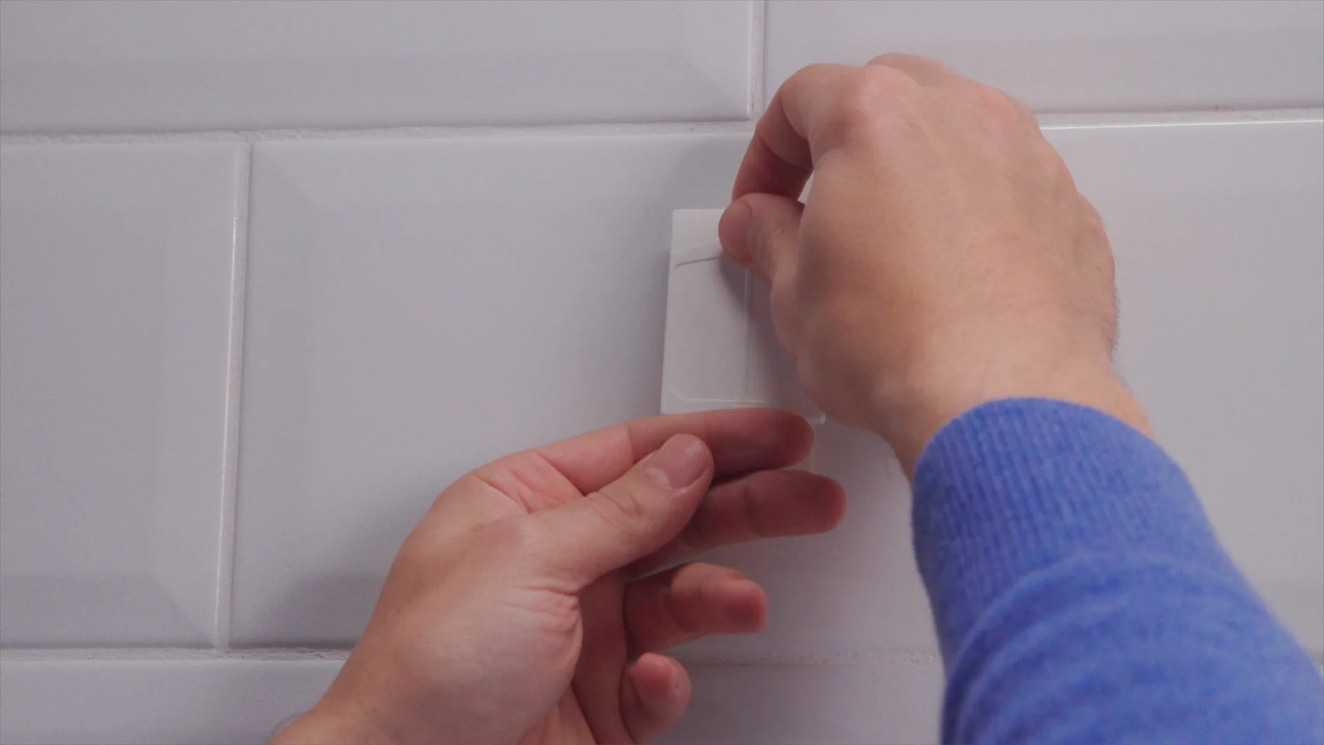 A person in a blue sweater applies a white adhesive hook to a tiled wall using tesa tape. (This text has been generated by AI)