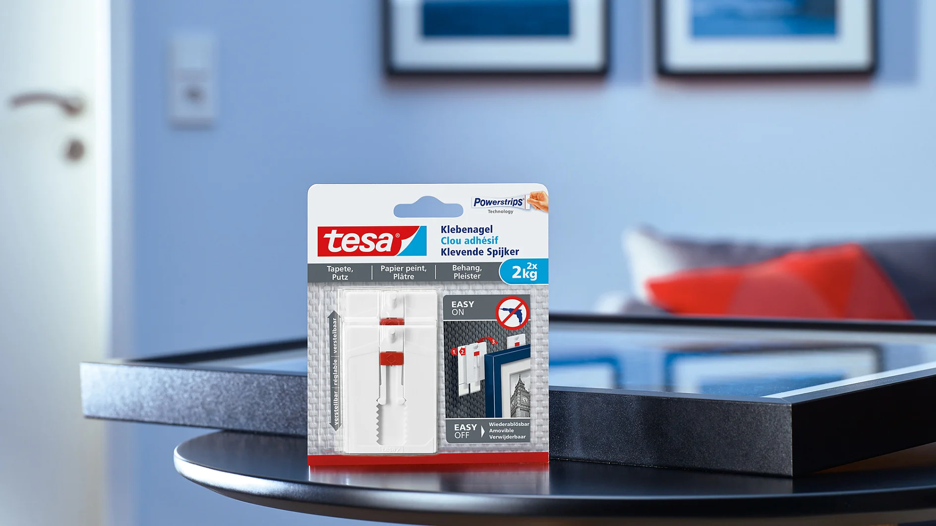 A package of tesa tape adhesive nails is placed on a black round table. Behind the table are two framed pictures on a blue wall and a partially open white door. The packaging shows installation instructions and a weight capacity of 2 kg. (This text has been generated by AI)