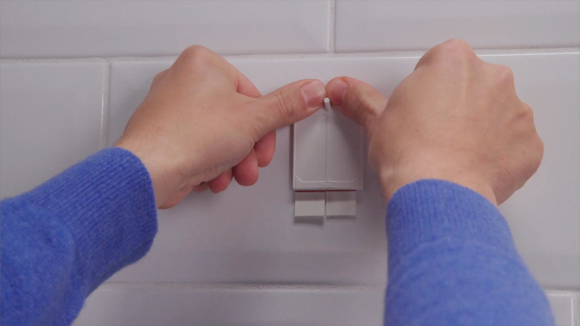 Two hands press a rectangular adhesive hook with a white strip against a white tiled wall using tesa tape. The person is wearing a blue long-sleeve shirt. The hook appears to be in the process of being secured to the surface. (This text has been generated by AI)