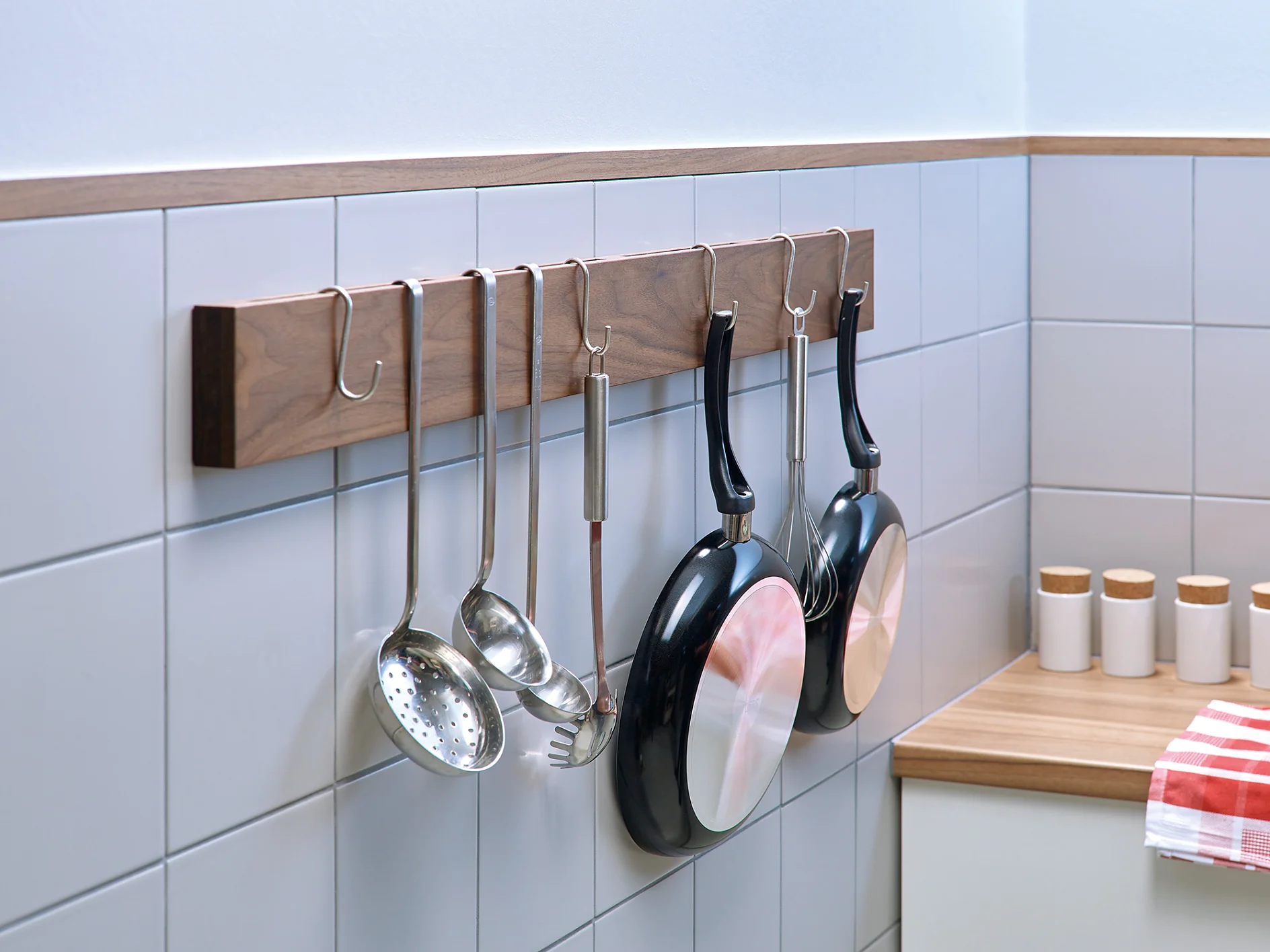 A kitchen wall features a wooden rack holding two frying pans, a ladle, a slotted spoon, and a pasta fork. Five small containers with wooden lids sit on the countertop nearby. A red and white checkered cloth is partially visible. (This text has been generated by AI)