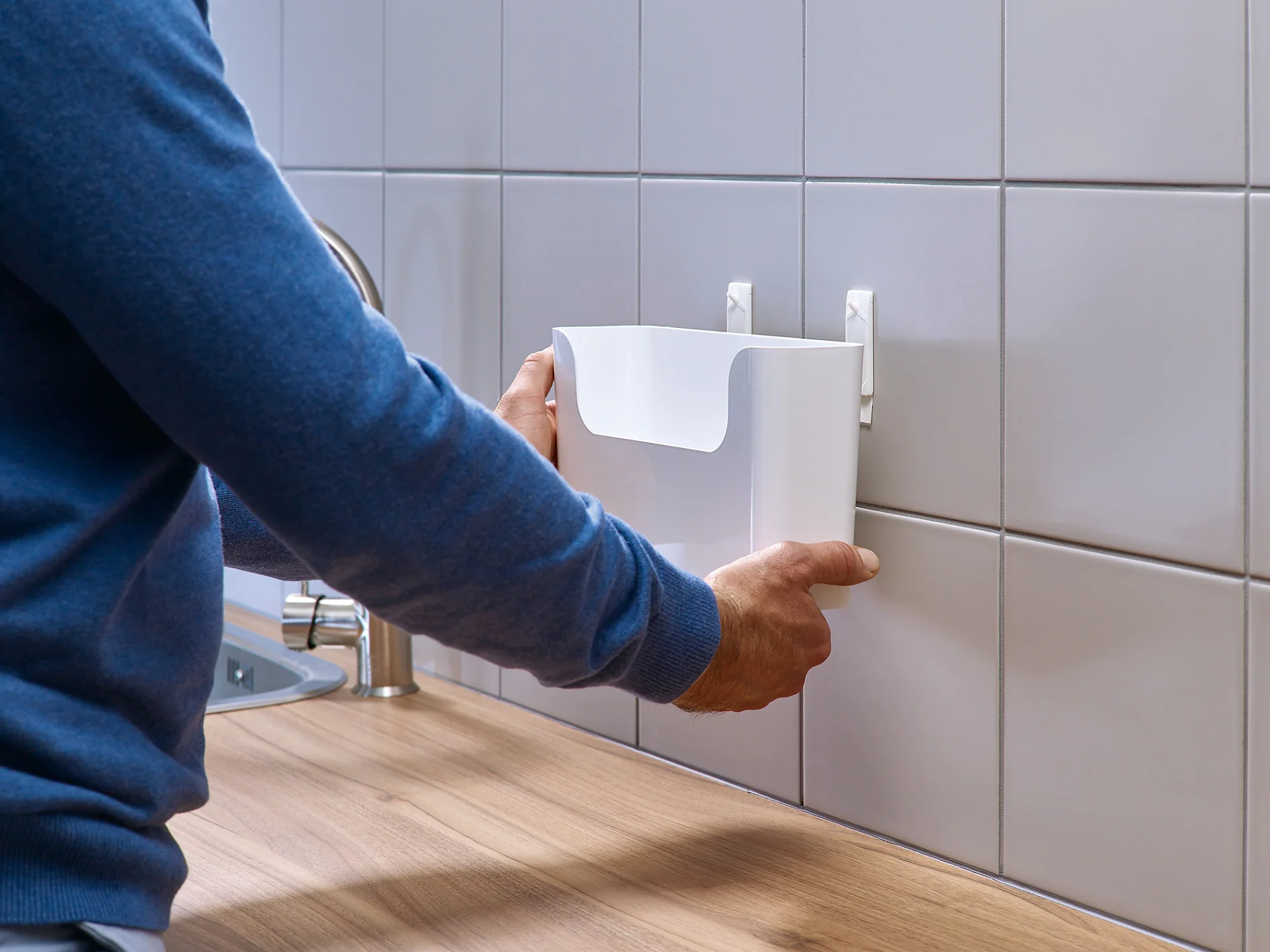 A person wearing a blue sweater is attaching a white plastic holder to hooks on a tiled kitchen wall using tesa tape. The wooden countertop and stainless steel sink are partially visible. (This text has been generated by AI)
