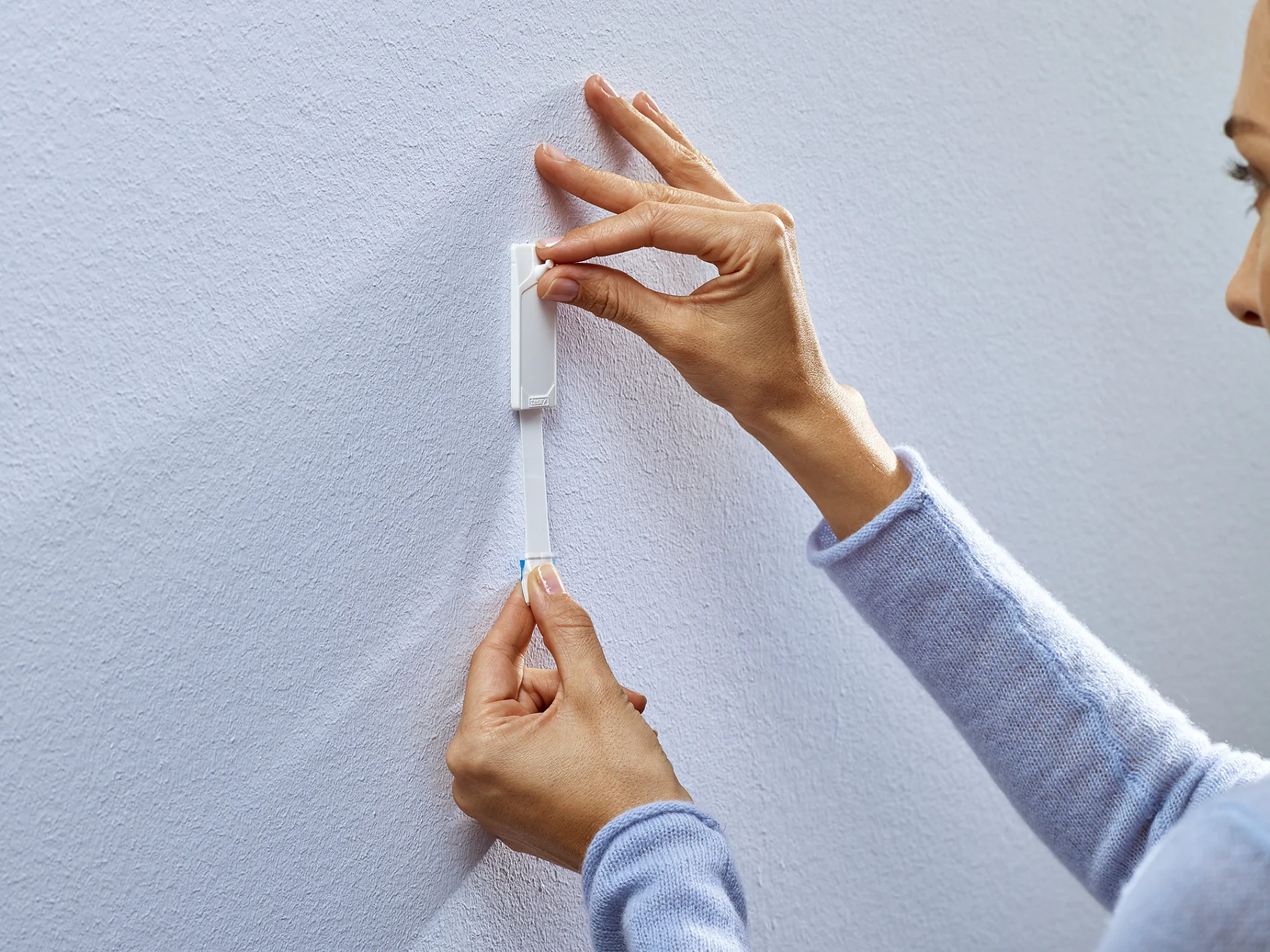 A person wearing a light blue sweater is using a measuring tesa tape to mark a spot on a light gray wall. They are holding the tesa tape with one hand and positioning the marker with the other. (This text has been generated by AI)