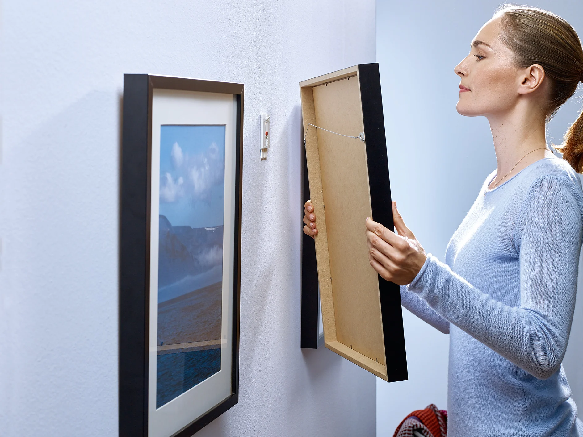 A woman with her hair tied back is hanging a framed picture on a white wall. She holds the frame with both hands next to an already hung picture of a landscape. The room is well-lit, and she seems focused on her task, using tesa tape for secure placement. (This text has been generated by AI)