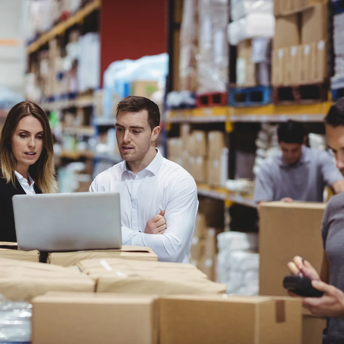 A group of warehouse workers stand by a table filled with packages. Two are engaged with a laptop, while another tallies items using a handheld device. Shelves stocked with boxes and supplies, including tesa tape, are visible in the background. (This text has been generated by AI)