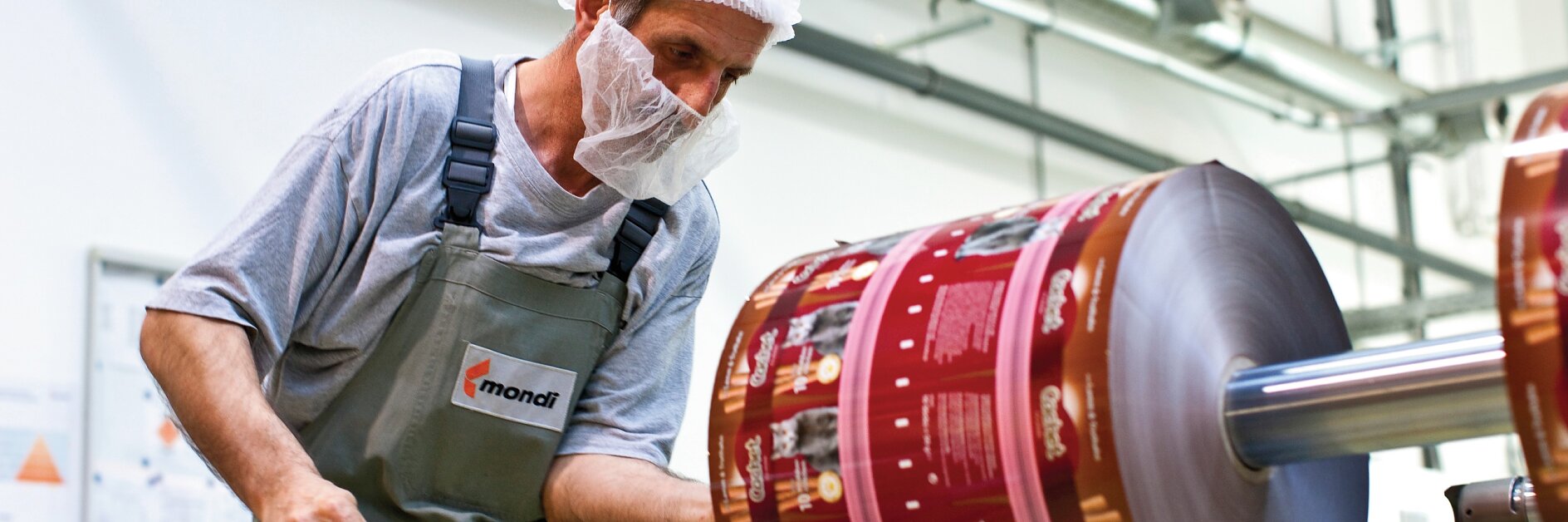 A worker wearing a hairnet and face mask inspects a large roll of printed packaging material in a factory setting. He holds a tool and is dressed in a gray shirt and green overalls. Machinery, tesa tape, and industrial elements are visible in the background.