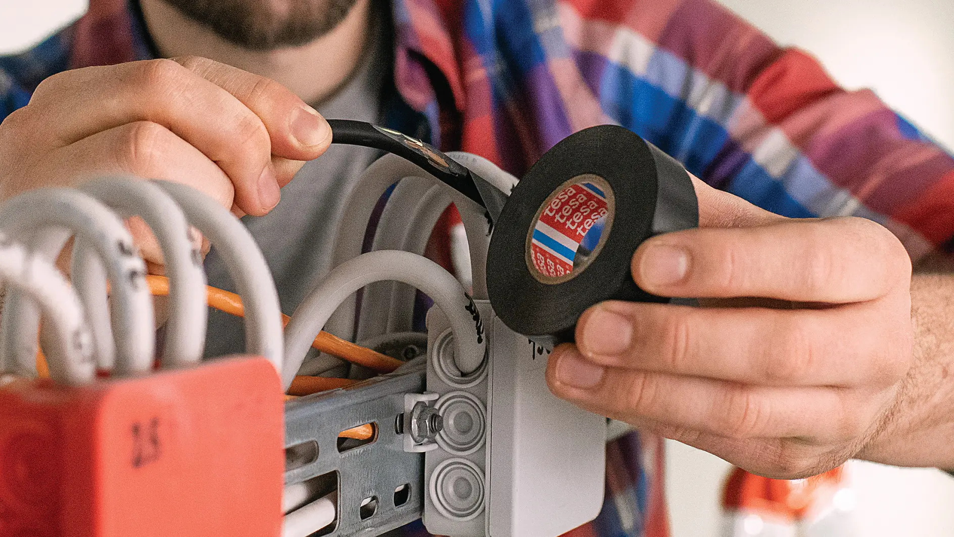 A person wearing a plaid shirt is holding a roll of black tesa tape and is applying it to wires in an electrical box. The scene appears to involve electrical work or installation, with multiple cables being organized.