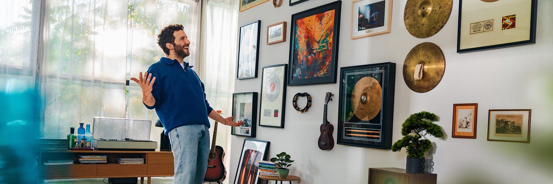 Un homme portant un pull bleu se tient dans une pièce décorée de disques en or encadrés, de guitares et dœuvres dart sur les murs. Il y a un tourne-disque et des haut-parleurs. La pièce est dotée de grandes fenêtres et de plantes en pot. (This text has been generated by AI)