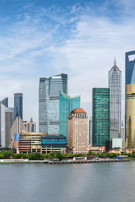 Panoramic view of Shanghais skyline featuring the Oriental Pearl Tower and various skyscrapers along the waterfront, under a clear blue sky. The Huangpu River is in the foreground with boats visible on the water. (This text has been generated by AI)