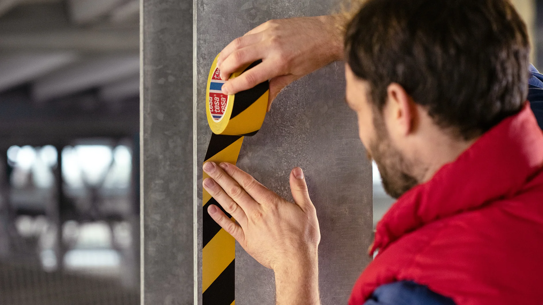 Une personne applique verticalement du ruban tesa de signalisation de danger noir et jaune sur un pilier en béton gris dans ce qui semble être un garage de stationnement. Seuls les mains et le profil de la personne sont visibles. (This text has been generated by AI)