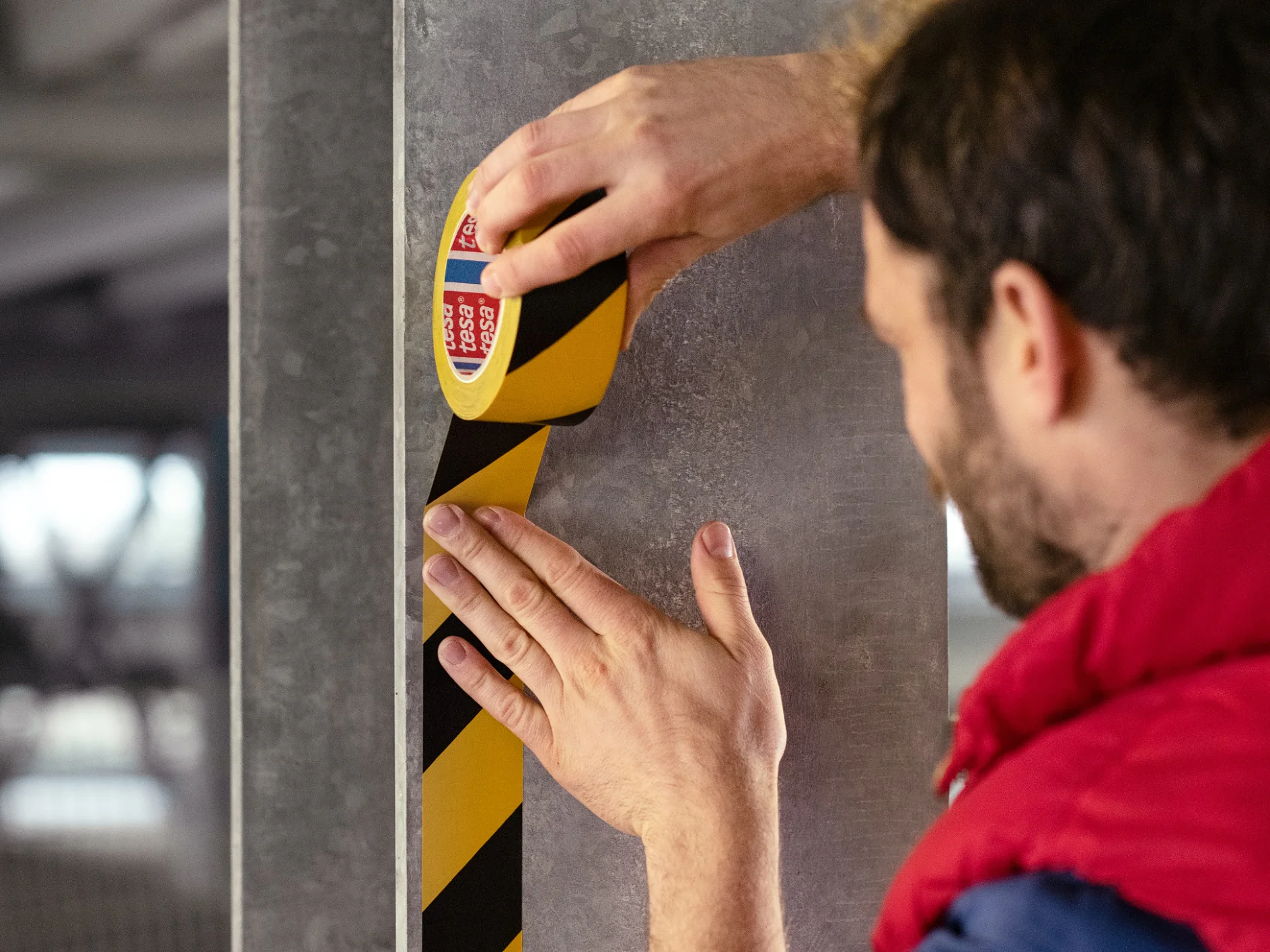 Une personne applique verticalement du ruban tesa de signalisation de danger noir et jaune sur un pilier en béton gris dans ce qui semble être un garage de stationnement. Seuls les mains et le profil de la personne sont visibles. (This text has been generated by AI)