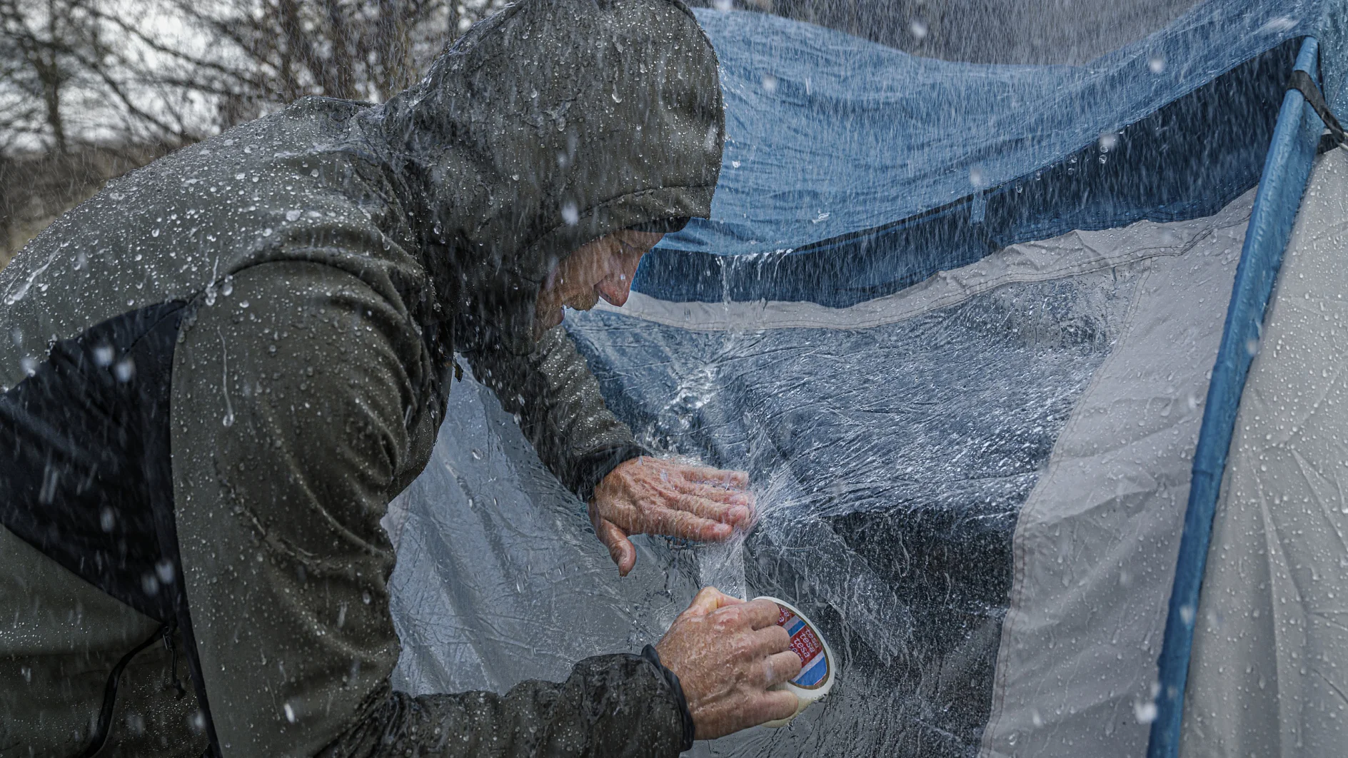 Une personne vêtue d'une veste de pluie applique du ruban adhésif tesa sur la couture d'une tente alors qu'il pleut abondamment. L'eau tombe à verse et la toile de la tente est visiblement mouillée. Des arbres sans feuilles sont visibles à l'arrière-plan. (This text has been generated by AI)