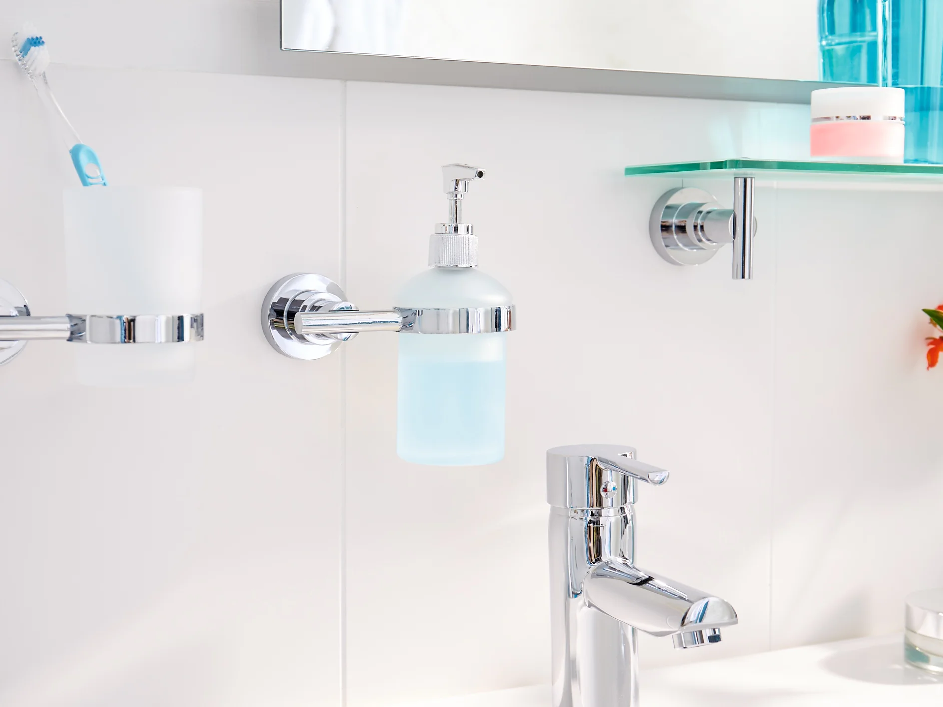 In the bathroom sink area, a chrome faucet complements a white counter. A toothbrush and holder, along with a soap dispenser, are securely mounted using tesa tape on the tiled wall. A shelf holding toiletries is also affixed with tesa tape. On the right side of the counter sits a small blue vase filled with orange flowers. (This text has been generated by AI)