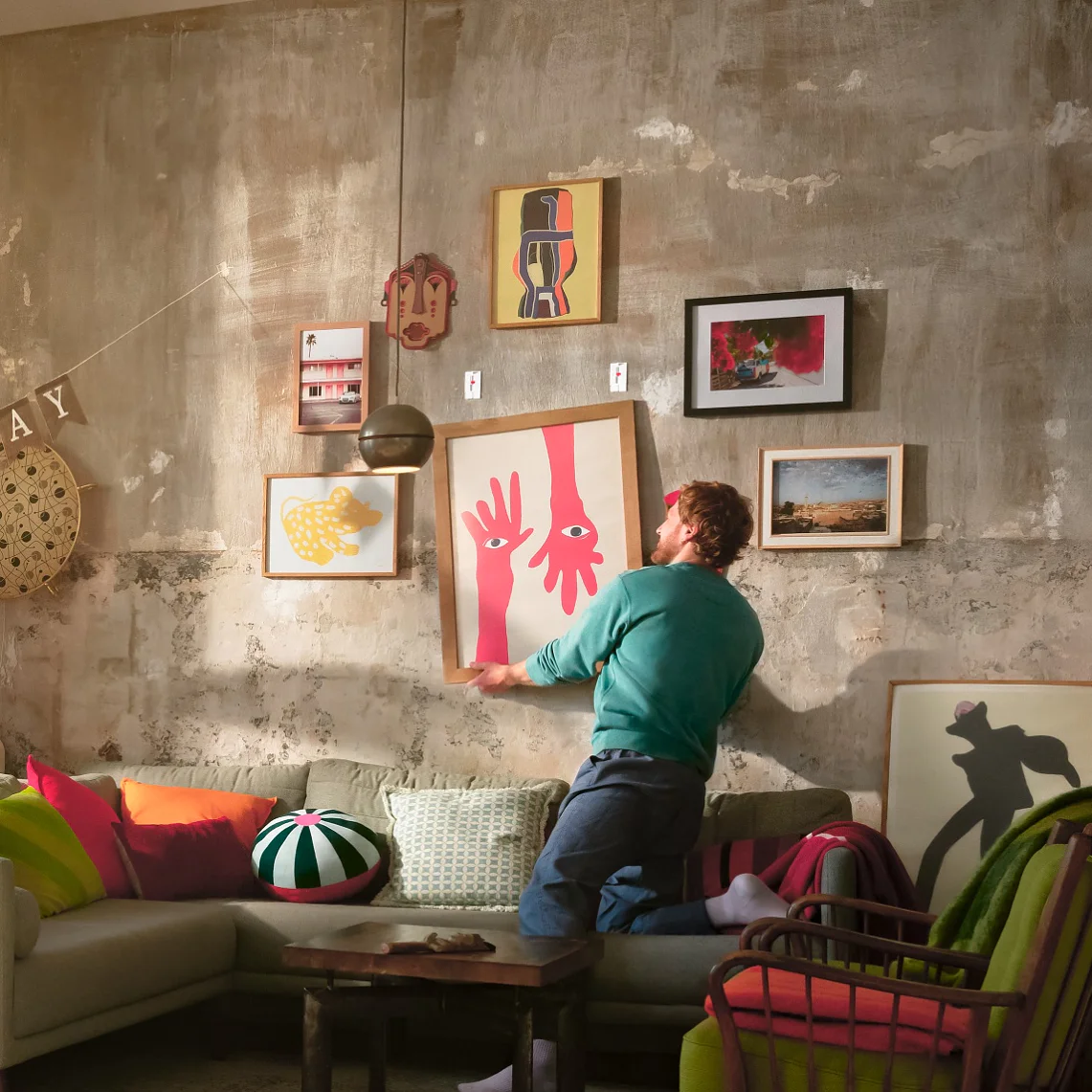 Une personne se tient dans un salon coloré décoré d'une banderole "Happy Birthday" et arrange des œuvres d'art sur un mur à l'aide de ruban adhésif tesa. L'espace est agrémenté de plantes, d'étagères, d'un canapé sectionnel gris et de divers coussins. (This text has been generated by AI)