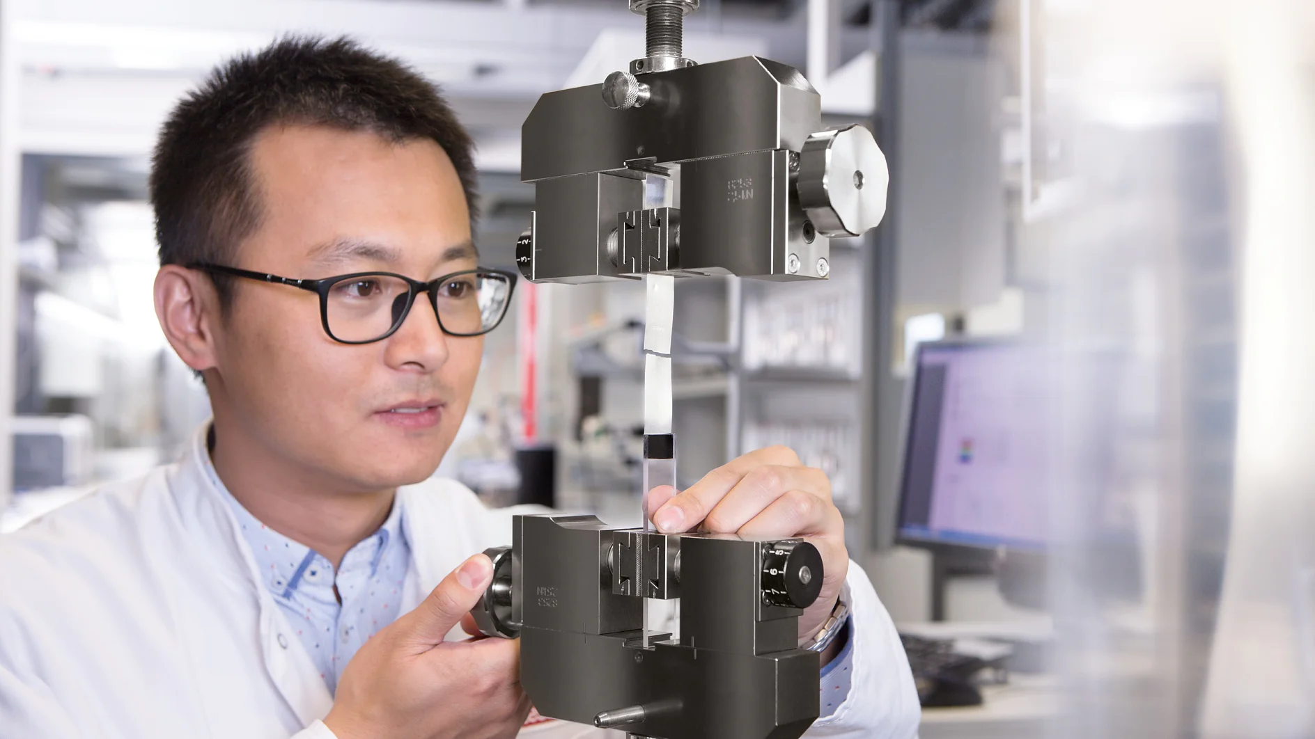 Une personne portant des lunettes et une blouse de laboratoire ajuste un échantillon de matériau fixé avec du ruban adhésif tesa dans une machine d'essai mécanique dans un laboratoire, avec des écrans d'ordinateur et des équipements de laboratoire visibles à l'arrière-plan. (This text has been generated by AI)