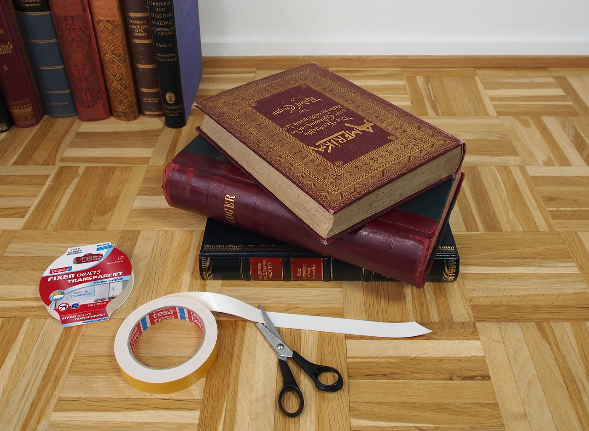 Une pile de trois gros livres repose sur un parquet en bois. À proximité, on peut voir un rouleau ouvert de ruban adhésif transparent tesa, des ciseaux et un paquet étiqueté pour du ruban adhésif tesa. Dautres livres sont debout à larrière-plan. (This text has been generated by AI)