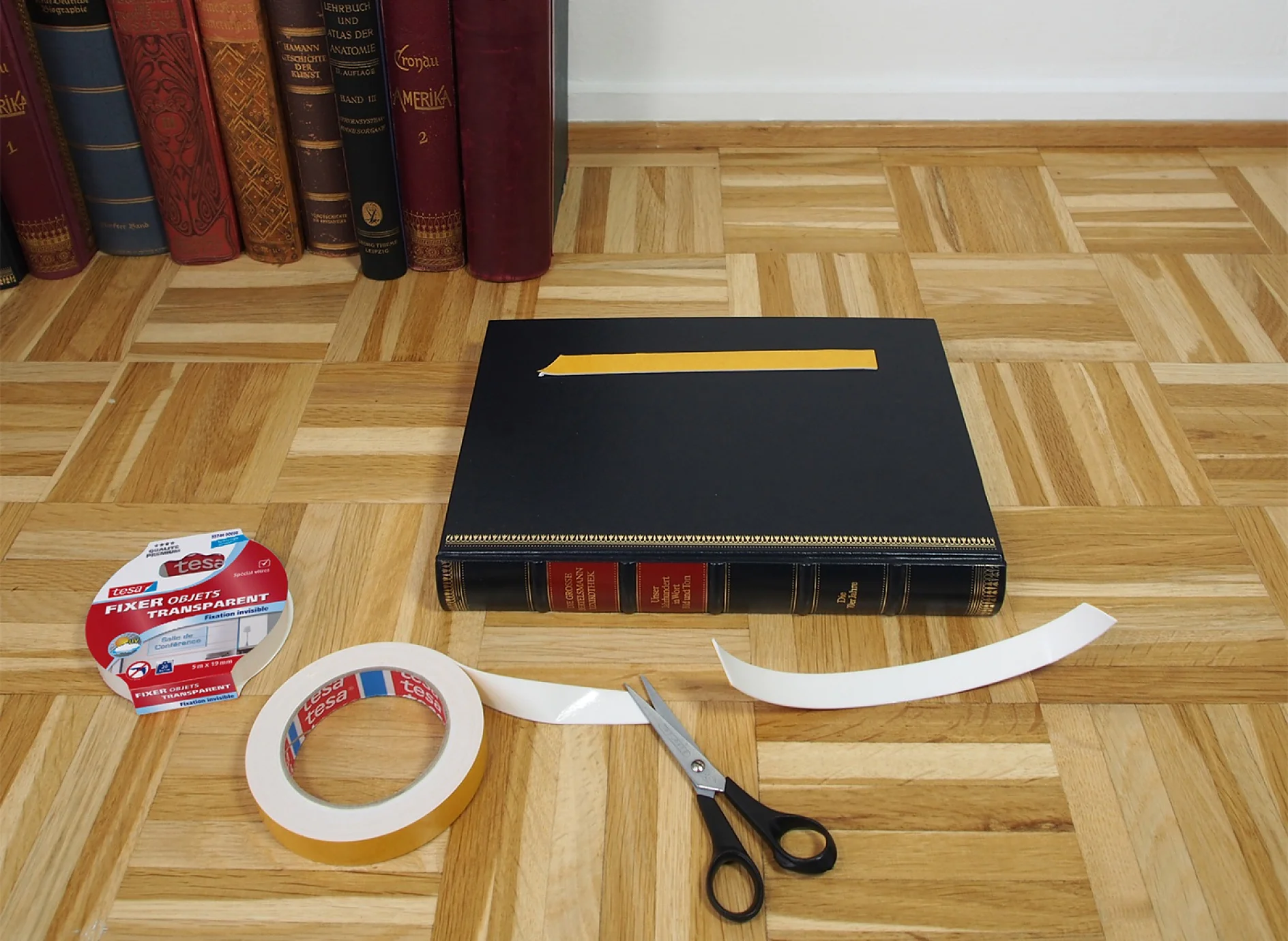 Un livre noir est posé sur un plancher en bois, avec une bande de ruban adhésif Tesa dessus. À proximité se trouvent des ciseaux et deux rouleaux de ruban adhésif Tesa transparent, dont lun est partiellement déroulé. Des livres sont debout contre un mur en arrière-plan. (This text has been generated by AI)