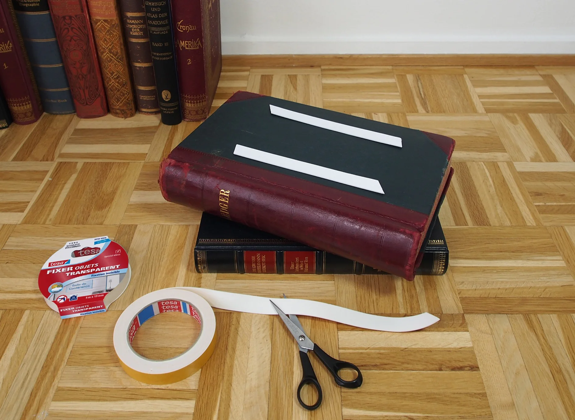 Deux livres à couverture rigide sont empilés sur un plancher en bois. Au-dessus de la pile, un grand livre est recouvert de deux bandes blanches. À proximité se trouvent des ciseaux, un rouleau de ruban adhésif Tesa, des bandes adhésives blanches et un paquet de ruban adhésif Tesa transparent. Des étagères sont à larrière-plan. (This text has been generated by AI)