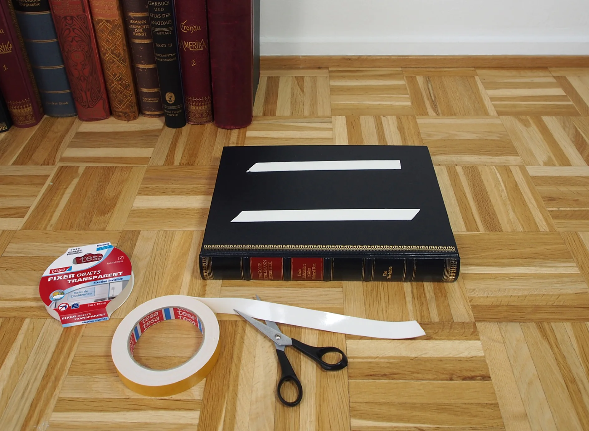 Un livre noir avec deux bandes blanches sur sa couverture est posé sur un plancher en bois. À proximité se trouvent un rouleau de ruban adhésif tesa transparent, une paire de ciseaux noirs et un distributeur de ruban adhésif tesa vide. Une rangée de livres colorés est visible en arrière-plan. (This text has been generated by AI)