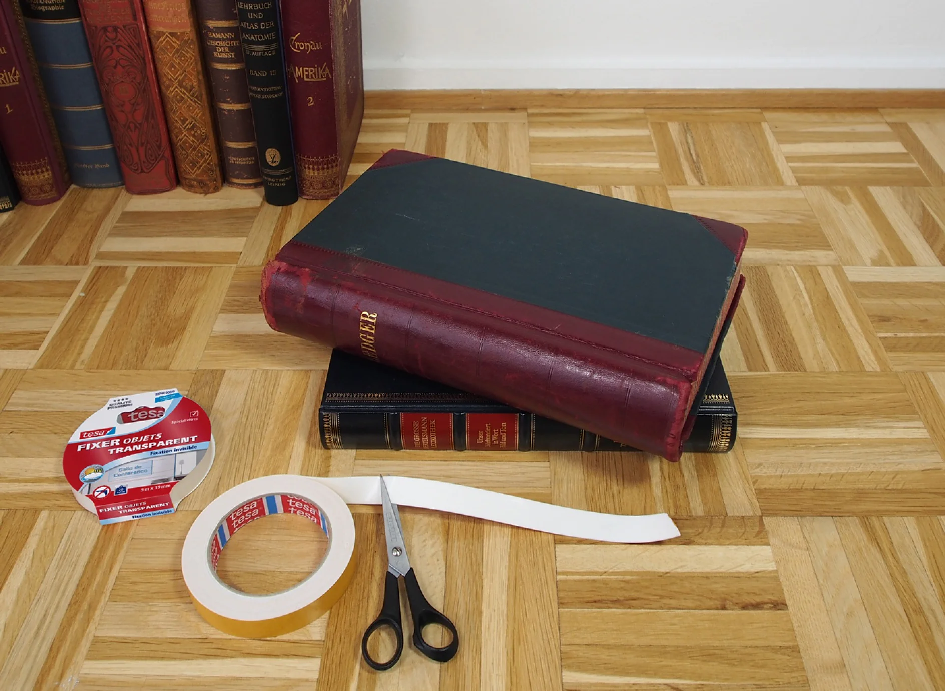 Un grand livre fermé à la couverture sombre et au dos rouge est posé sur deux autres livres empilés. À proximité se trouvent deux rouleaux de ruban adhésif tesa, une paire de ciseaux à manche noir et une bande de tissu blanc, le tout sur un parquet en bois. (This text has been generated by AI)