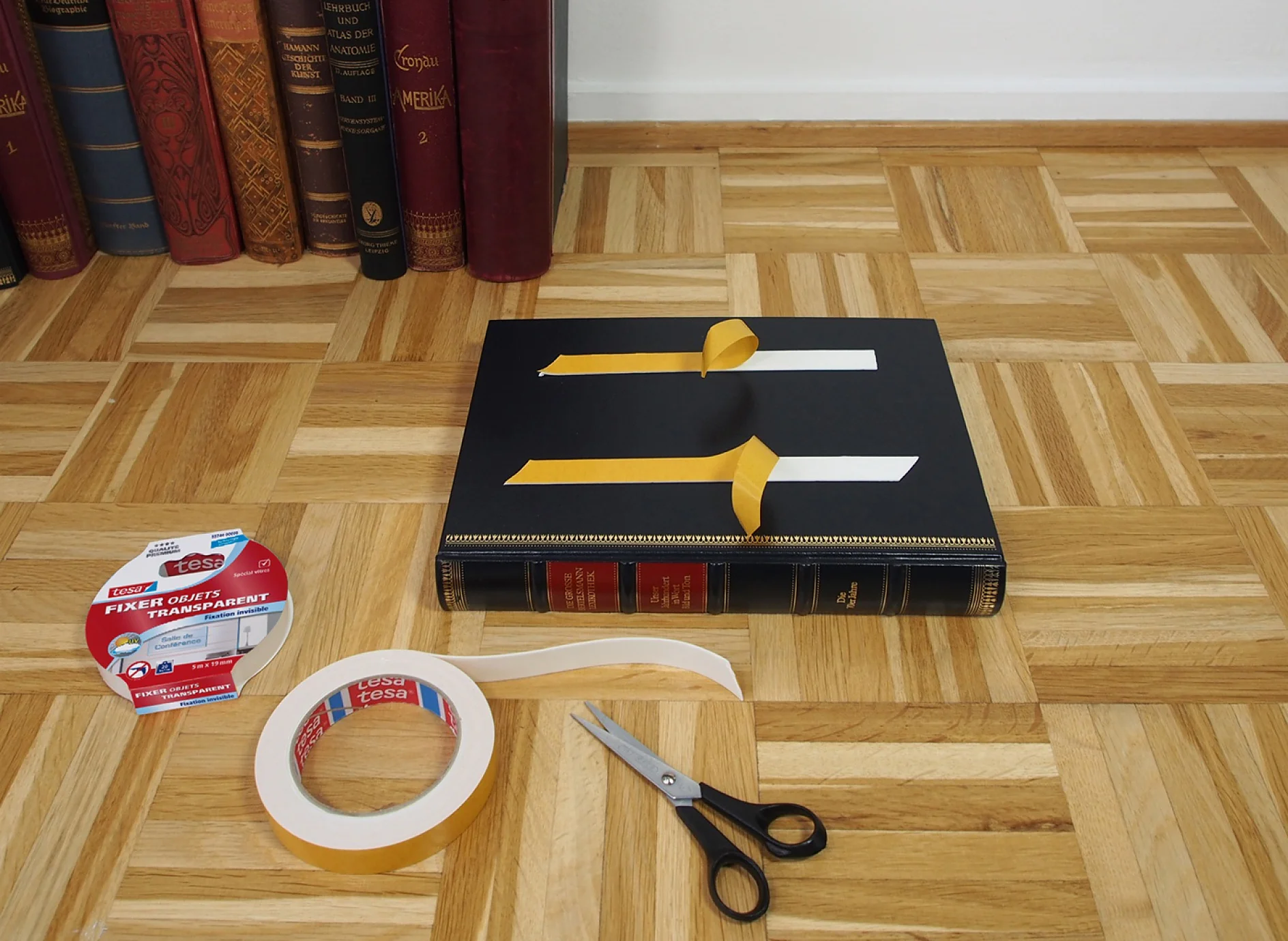 Un livre à couverture rigide est posé sur un plancher en bois avec deux morceaux de ruban adhésif tesa dépassant de la couverture. À proximité se trouvent une paire de ciseaux, un rouleau de ruban adhésif tesa transparent et son emballage. Une rangée de livres est visible en arrière-plan. (This text has been generated by AI)
