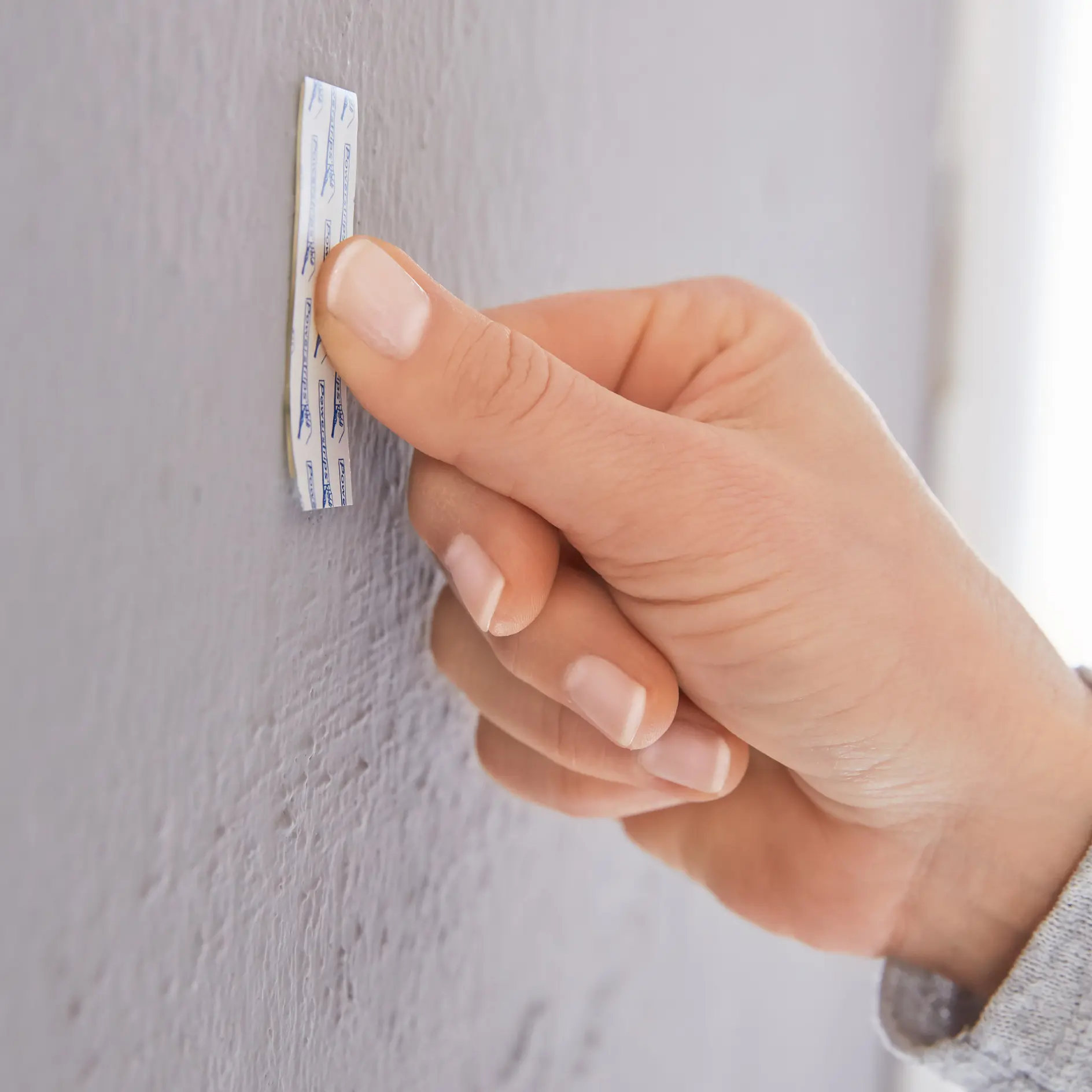 A persons hand is sticking a small tesa tape strip onto a textured gray wall. The tesa tape strip has a blue chevron pattern and a white backing. The persons thumb presses firmly on the tape to adhere it to the wall surface. (This text has been generated by AI)