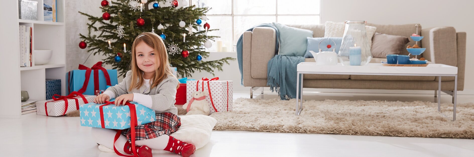 Une jeune fille aux cheveux longs est assise sur un coussin blanc dans un salon, tenant un cadeau bleu emballé avec du ruban adhésif rouge. Un sapin de Noël décoré avec des cadeaux se trouve à proximité. La pièce comprend un canapé beige, une table basse blanche et une décoration lumineuse. Une lumière naturelle éclatante remplit lespace. (This text has been generated by AI)