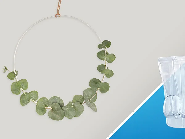 A decorative hoop with green eucalyptus leaves is displayed against a light background. Beside it, there is a transparent tesa tape wall hook on a blue surface. (This text has been generated by AI)