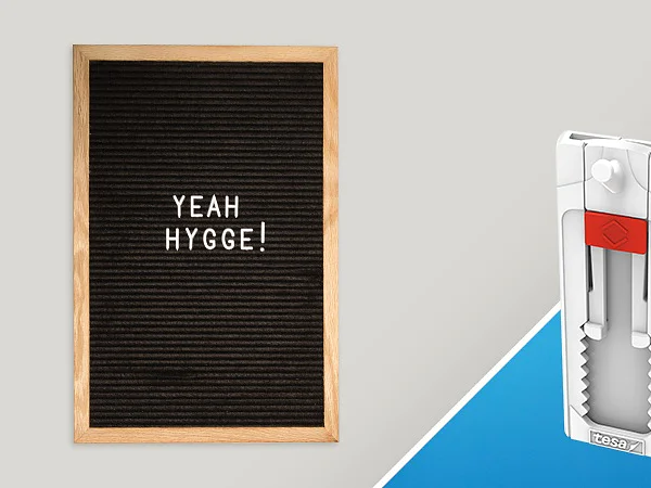 A black felt letter board with the words YEAH HYGGE! in white letters is on the left. On the right, there is a white and red craft tool featuring tesa tape with a grooved design on a blue background. (This text has been generated by AI)