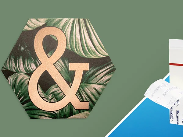 A hexagonal wall art piece with a large gold ampersand symbol in front of green leaves is set against a green background. Beside it, on a blue and white diagonal background, are tesa tape strips and their packaging. (This text has been generated by AI)