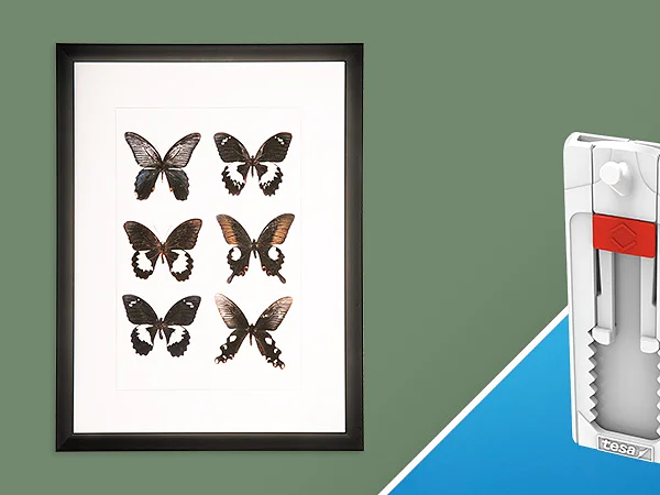 A framed display with nine brown butterflies is on a green background. Next to it, on a blue triangular background, is a white and red wall-hanging tool with a serrated grip and hook at the top, secured with tesa tape. (This text has been generated by AI)