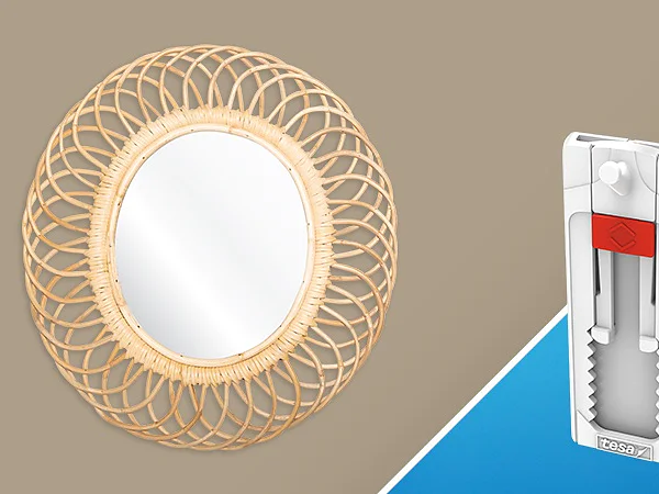 A round mirror with a decorative rattan frame is on the left against a beige background. On the right, there is a white plastic adhesive tool featuring tesa tape with a red component against a blue background. (This text has been generated by AI)