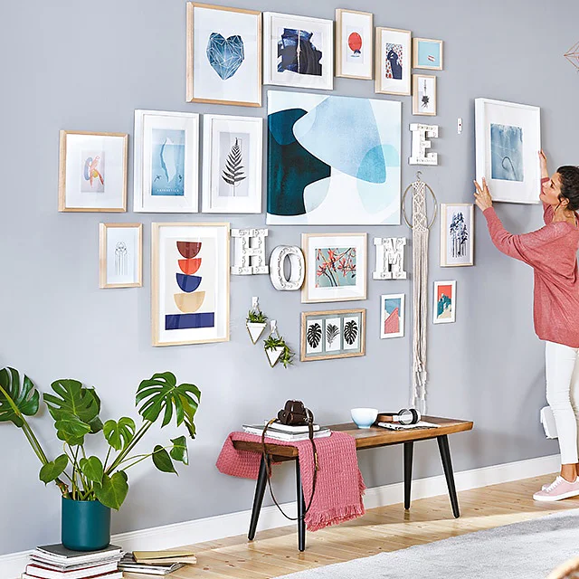 Une femme en haut rose et pantalon blanc accroche un tableau encadré sur un mur gris rempli dœuvres dart diverses et dimpressions encadrées à laide de ruban adhésif tesa. La pièce comprend du parquet, des plantes, un banc en bois et des rideaux de couleur claire près dune fenêtre. (This text has been generated by AI)