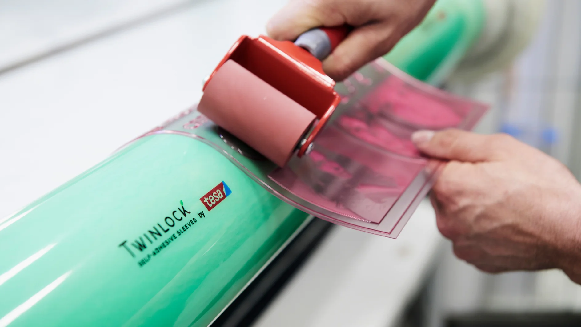 Close-up of hands using a small roller to apply a pink sheet onto a large green cylinder labeled Twinlock. The cylinder also displays the phrase Self Adhesive Sleeves, with tesa tape being used in the process. (This text has been generated by AI)