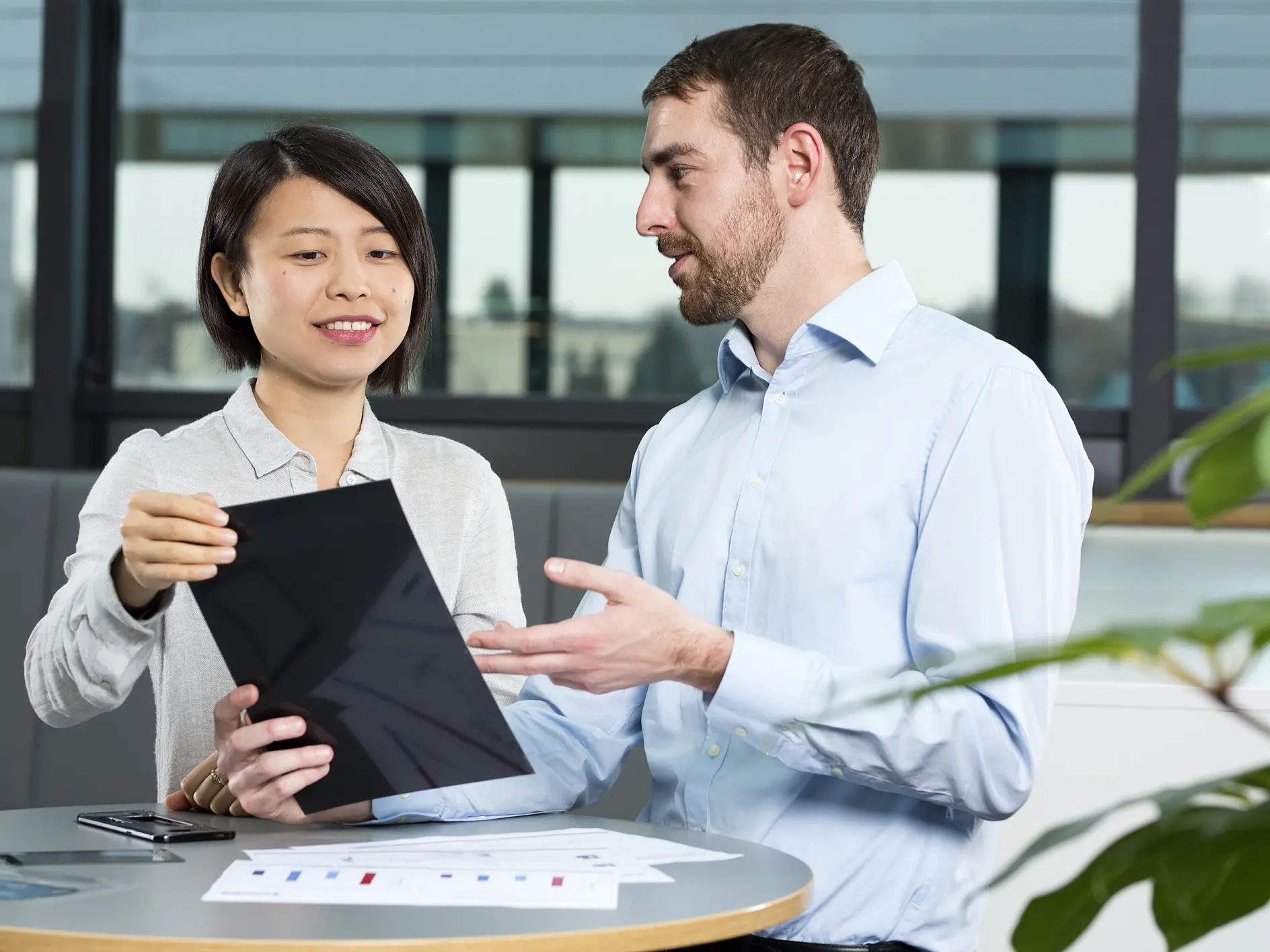 Two people are discussing something on a tablet in an office setting. The woman is holding the tablet, while the man gestures toward it. A document with graphs is on the table secured with tesa tape. They are seated near a window with a view outside. (This text has been generated by AI)