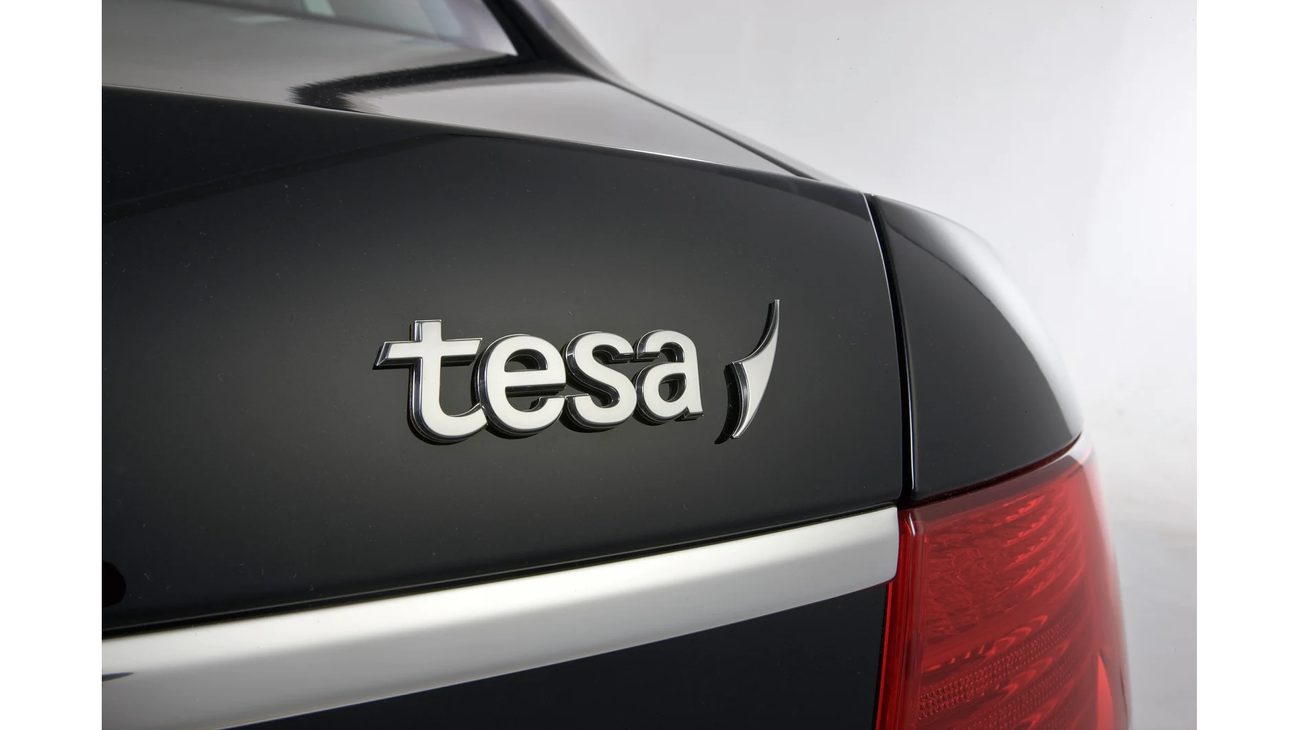 Close-up of the rear section of a black car. The logo tesa is prominently displayed on the trunk. The right taillight is partially visible, along with a silver trim along the edge of the trunk. (This text has been generated by AI)