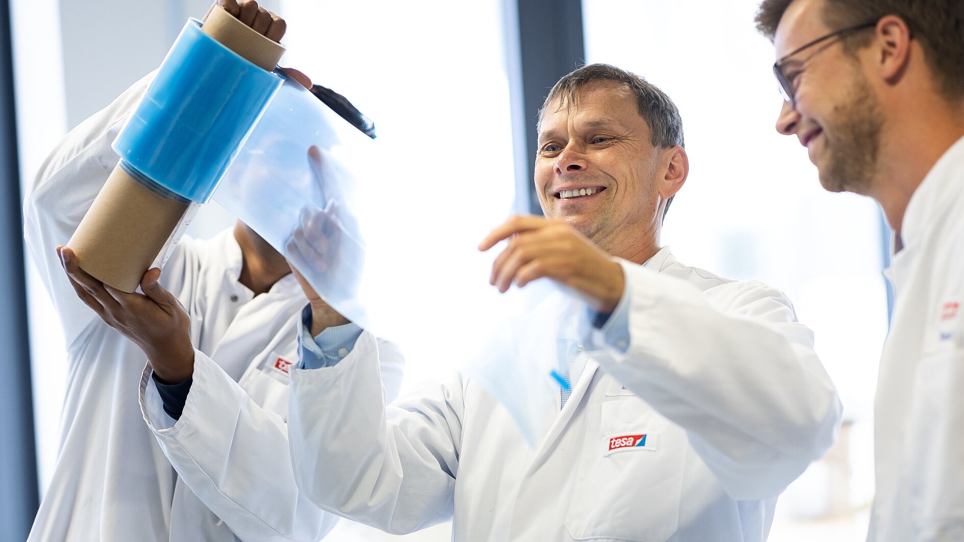 Three people in lab coats examine a roll of transparent blue tesa tape in a bright laboratory setting. One person holds the tesa tape upright, while another pulls a section away. They appear focused and engaged. (This text has been generated by AI)